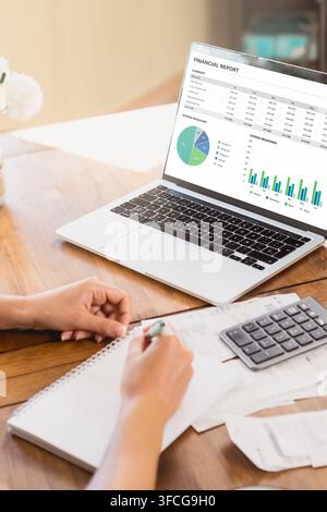 Overhead view of hands taking notes beside a laptop displaying financial charts, calculator and documents on a wooden home office desk. Stock Photo