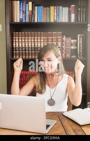 Happy young woman with glasses raises both fists and celebrates good news on her laptop at a wooden home office desk. Stock Photo