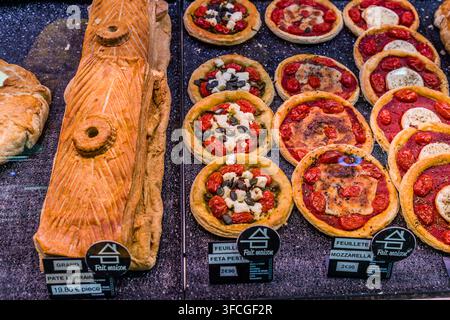 A large pâté Lorrain and round puff-pastry tartlets topped with tomatoes, olives, feta and mozzarella lie in the display case of a bakery on Place Charles III in Nancy, Grand Est, France, labelled "Feuilleté Feta Pesto" and "Feuilleté Mozzarella" on the signage. Pâté Lorrain is a puff-pastry pie from Lorraine filled with pork and veal. Stock Photo