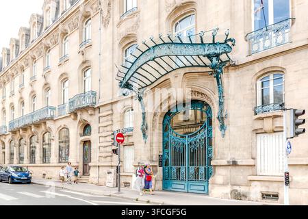 A curved Art Nouveau canopy of glass and wrought iron spans the entrance of the Chambre de Commerce et d'Industrie de Meurthe-et-Moselle on Rue Henri Poincaré in Nancy, Grand Est, France. Passers-by, including two women wearing sun hats, walk past the sandstone façade. Stock Photo
