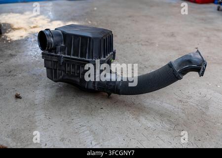 Black air filter housing rests on concrete floor with flexible intake hose attached. Dusty plastic body and clamp points suggest removed automotive Stock Photo