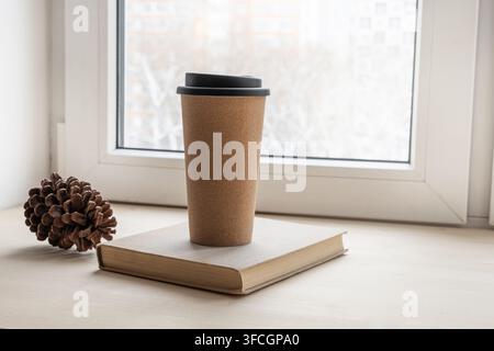 Reusable takeaway coffee cup on a book with pine cone by bright window, cozy minimalist home scene for coffee break, reading and autumn lifestyle. Stock Photo