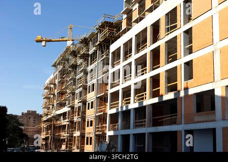 An unfinished multi-story concrete and brick building stands beside scaffolding and a tower crane under a clear blue sky. Bright sunlight highlights t Stock Photo