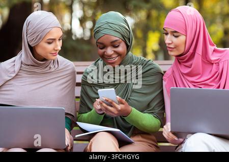 Muslim Lady With Smartphone Learning Online With Friends Using Laptops Sitting Together In University Campus Park Outdoor. Educational Application, Di Stock Photo