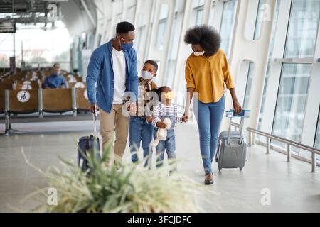 A Black family with two children wearing protective face masks and carrying suitcases walks through a modern airport terminal Stock Photo