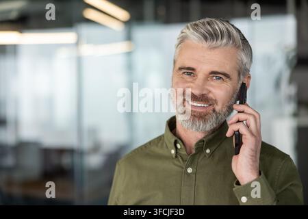 Confident mature man with gray hair and beard smiling, talking on a smartphone, representing success, business communication, and professional connection in a modern office Stock Photo