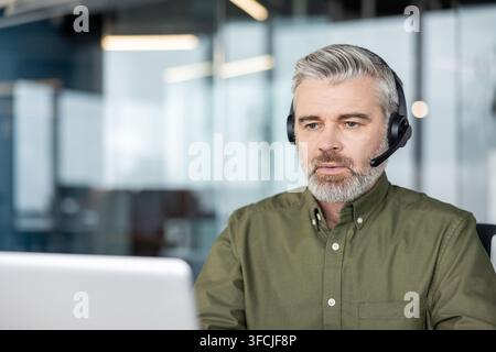 Mature man with a headset working in an office, focusing on his laptop screen while communicating with clients, representing dedicated professional service and technical assistance Stock Photo