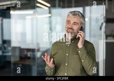 Mature businessman talking on a mobile phone, gesturing with his hand while having a conversation in a modern office environment, representing communication and business networking Stock Photo