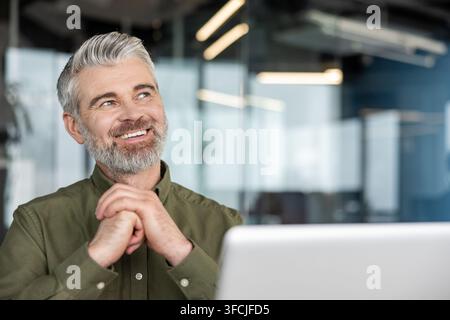 Successful senior businessman with gray hair and beard sitting in an office, looking up, smiling, and feeling inspired while generating new creative business ideas Stock Photo