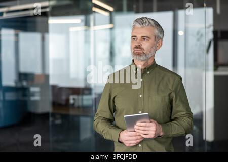 Mature businessman with gray hair and beard holding a digital tablet, looking away thoughtfully while standing in a modern office with glass walls, reflecting leadership and strategic thinking Stock Photo