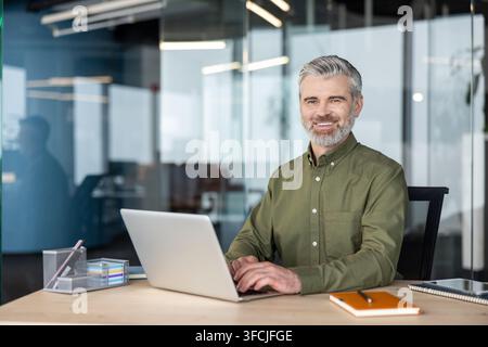 Mature businessman with gray hair and beard sitting at a wooden desk, working on a laptop in a modern office, portraying success, professionalism, and confidence Stock Photo