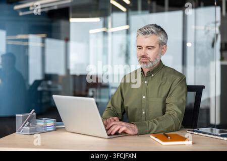 Experienced professional man sitting at a wooden desk, focused on typing on a laptop computer in a contemporary corporate office environment, highlighting business productivity Stock Photo