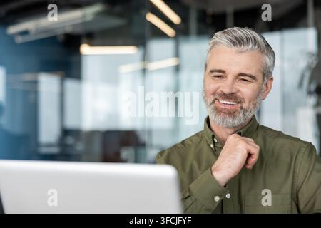 Senior gray-haired, bearded businessman smiling confidently at his laptop, working productively in a modern office with glass walls, focused and engaged in a video call Stock Photo