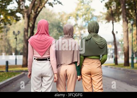 Back View Of Three Modern Muslim Women Walking Together Wearing Hijab Headscarves In City Park Outdoors. Islamic Females Friendship And Arabic Fashion Stock Photo