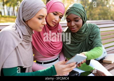 Islamic Females Students Using Smartphone With New Educational Application Learning Together Sitting On Bench In Campus Park. People And Gadgets, Mode Stock Photo