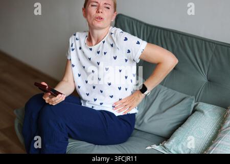 Sick woman with stomach pain using AI chatbot on smartphone for health advice while resting on sofa at home. Stock Photo