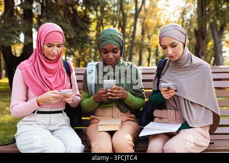 Group Of Diverse Muslim Ladies Using Smartphones Texting And Browsing Internet Sitting On Bench Outdoors. People, Technology And Gadgets, New Mobile A Stock Photo