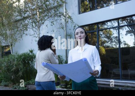 Diverse female coworkers holding blueprints, in white blouse, talking on smartphone by glass Stock Photo