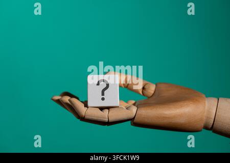 Wooden mannequin hand holding a small white wooden block with printed question mark symbol against turquoise background, close up. Concept of question Stock Photo
