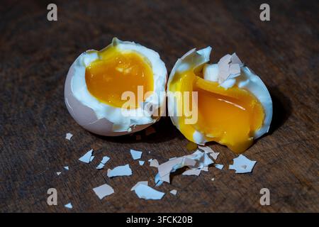 Soft boiled chicken egg cracked open into two halves showing bright runny orange yolk and white albumen on dark rustic wooden background with pieces o Stock Photo