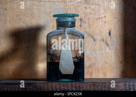 Vintage apothecary glass bottle with blank label on wooden shelf. Antique clear glass medicine jar filled with dark liquid or powder. Old pharmaceutic Stock Photo