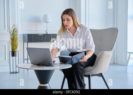 Focused businesswoman with notebook working on laptop in modern office space Stock Photo