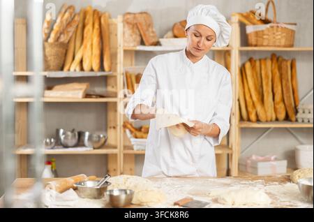 While woman working at bakery, forms dough, holds flat and thin piece in hands, stretches dough with hands Stock Photo