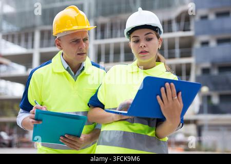 Two qualified engineers working on a construction site are discussing an important project, holding a laptop and an estimate ..in their hands Stock Photo