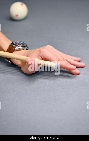A vertical studio photograph capturing a close-up of a person's hand forming a bridge on a pool table surface while holding a cue stick, with a white billiard ball in the background. Stock Photo