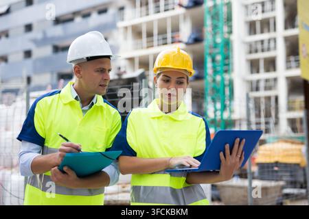 Two qualified engineers working on a construction site are discussing an important project, holding a laptop and an estimate ..in their hands Stock Photo