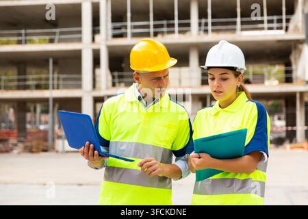 Two qualified engineers working on a construction site are discussing an important project, holding a laptop and an estimate ..in their hands Stock Photo