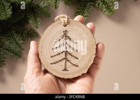 Christmas toy made of decorative wooden slice with ornament as Xmas tree on beige background. Handmade hobby. DIY. Nature material. Xmas decoration. Eco friendly. Zero waste. Sustainable gifts. View from above. Stock Photo
