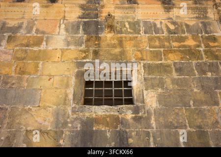 Old weathered stone wall with a small iron-barred rectangular window and a small stone gargoyle near the top edge. Stock Photo