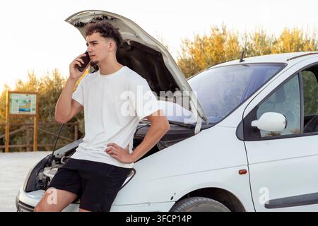 Young man using a smartphone, calling for help with his broken-down car, experiencing vehicle engine trouble Stock Photo
