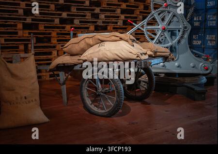 Vintage wooden hand truck loaded with burlap sacks of raw materials next to a historic port crane and stacked wooden pallets. Stock Photo