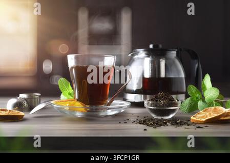 Glass cup and teapot full of tea on a wooden bench with a bowl of dried tea leaves and a kitchen in the background. Front view. Stock Photo