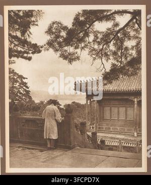 Unknown person at a house overlooking a lake, page, photomechanical print, photographer: Donald Mennie, Beijing, cliché maker: anonymous, c. 1915 - in or before 1920, paper, rotogravure, height 238 mm x width 203 mm, historical persons, landscapes with waters, waterscapes, seascapes (in the temperate zone), rural housing, eg country-house, villa, cottage, China., A sepia-toned scene shows a person wearing a light-colored long coat standing with their back to the camera, leaning on a carved stone balustrade and looking outward; to the right a traditional tiled roof with ornamental ridge figures Stock Photo
