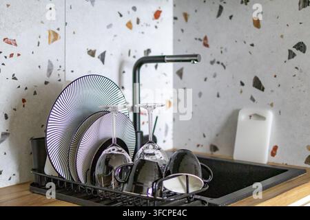 Clean dishes dry on rack beside sink after morning kitchen cleanup at home. Concept of household cleanliness and tidy kitchen maintenance during domes Stock Photo