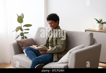 Cheerful black male student sitting on couch with book, reading and enjoying home studies. Young African American guy with textbook preparing for exam Stock Photo