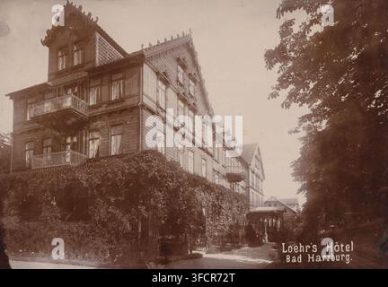 Loehr's Hôtel Bad Harzburg, Loehr's Hotel in Bad Harzburg, photograph, Bad Harzburg, 1890 - 1910, photographer, Lower Saxony, paper, pasteboard, baryta paper, daylight collodion silver print, height 149 mm x width 187 mm, hotel, hostelry, inn., A multi-story wooden building with decorative trim and patterned roof tiles fills the left and center of the scene, featuring several windows with curtains, ornate balconies, and carved gable details; dense climbing greenery covers the lower facade and spills over a hedge in front; a courtyard area with scattered chairs and a streetlamp sits near the Stock Photo