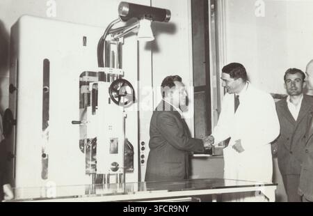 Palmiro Togliatti with surgeon Pietro Valdoni after donating medical equipment to a hospital in Rome, news photograph, inscription: 'Rome-On the first anniversary of the attempt at his life, ', Rome, publisher: International News Photos, United States of America, 1949-07, photographer, North America, paper, baryta paper, gelatin silver print, gelatin silver developing print, height 119 mm x width 183 mm, Person, Palmiro Togliatti, Pietro Valdoni, hospital, politics., A black and white photograph shows a room with a large medical or mechanical machine on the left featuring visible gears, wheels Stock Photo
