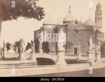 Padova, West facade of the Abbey of Santa Giustina in Padua, photograph, Padua, 1851 - 1900, photographer, pasteboard, paper, albumen print, albumendruk, height 179 mm x width 244 mm, height 270 mm x width 354 mm, sculpture, church (exterior), Basilica di Santa Giustina., A sepia-toned scene showing a row of stone statues mounted on pedestals along a balustraded bridge and riverside, with a large brick church building behind them featuring domed roofs and round windows, a tall bell tower on the right, and decorative architectural details; trees and foliage frame the upper left, and a person Stock Photo