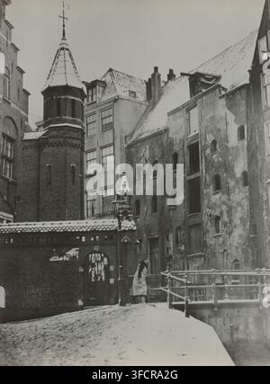 Snow-covered bridge and houses in the Spooksteeg in Amsterdam, Amsterdam, photograph, number: '587', 1910 - 1950, photographer, paper, baryta paper, gelatin silver print, developed gelatin silver print, height 166 mm x width 124 mm, urban housing, bridge in city across river, canal, Spooksteeg., A snowy urban scene showing several adjacent old buildings with varied facades and steep, tiled roofs dusted with snow; a prominent brick tower with a conical roof and a decorative finial stands near a lower brick wall topped with snow and an arched gate; next to the tower are multi‑story buildings wit Stock Photo