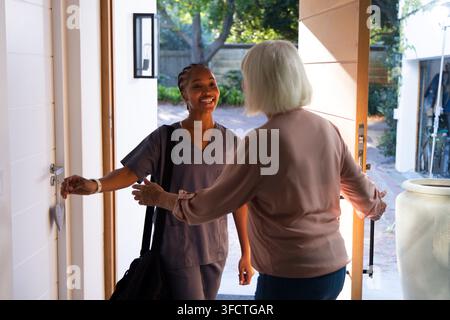 African American caregiver in scrubs greeting senior woman at door holding black bag and heart. Nurse, elderly, homecare, porch, sunlit, warmth, welco Stock Photo