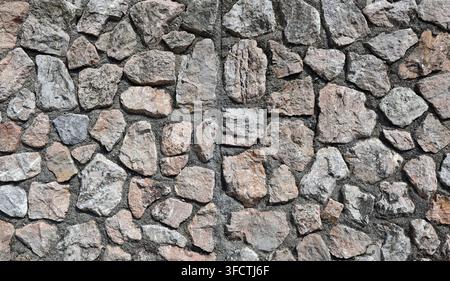 Stone wall made of gray and pinkish natural rocks on concrete base. Background and texture. Stock Photo