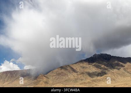 Dramatic storm clouds sweep over dry brown mountain ridges in a rugged highland landscape, with patches of blue sky and sunlight on the slopes. Stock Photo