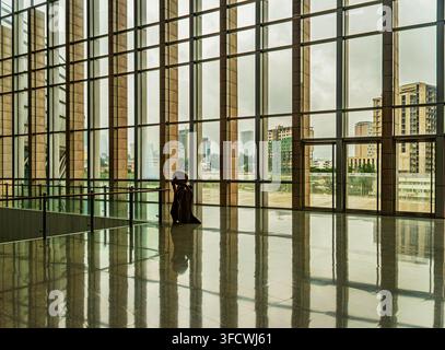 Solitary figure sits quietly in a vast, modern lobby with floor-to-ceiling glass walls. The person appears contemplative or tired, gazing outward at c Stock Photo
