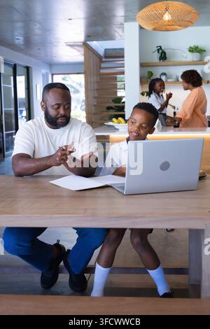 African American father and boy studying at wooden table using laptop, papers, pencil in kitchen Stock Photo