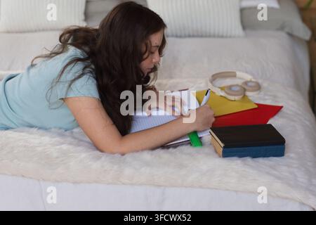 Teenage female lying on bed in bedroom studying, writing with pen and green ruler Stock Photo