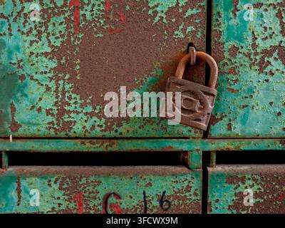 A locked, rusted steel padlock hanging on a weathered green metal mailbox with red number 8. Vintage architectural detail showing corrosion texture Stock Photo